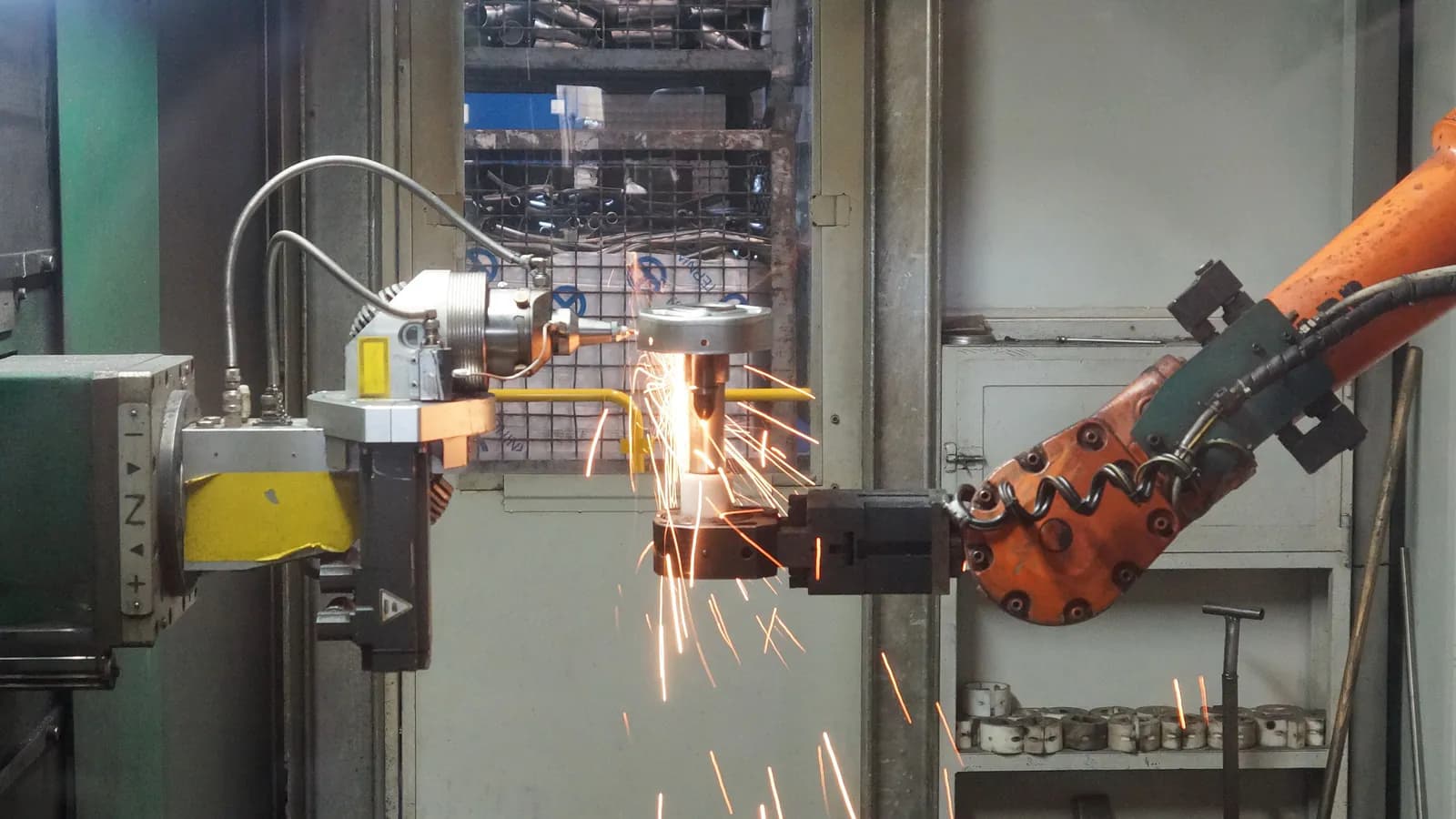 An industrial robotic arm cuts a metal part on a factory floor, sparks flying in every direction, while the surrounding equipment stays under an operator's watch.