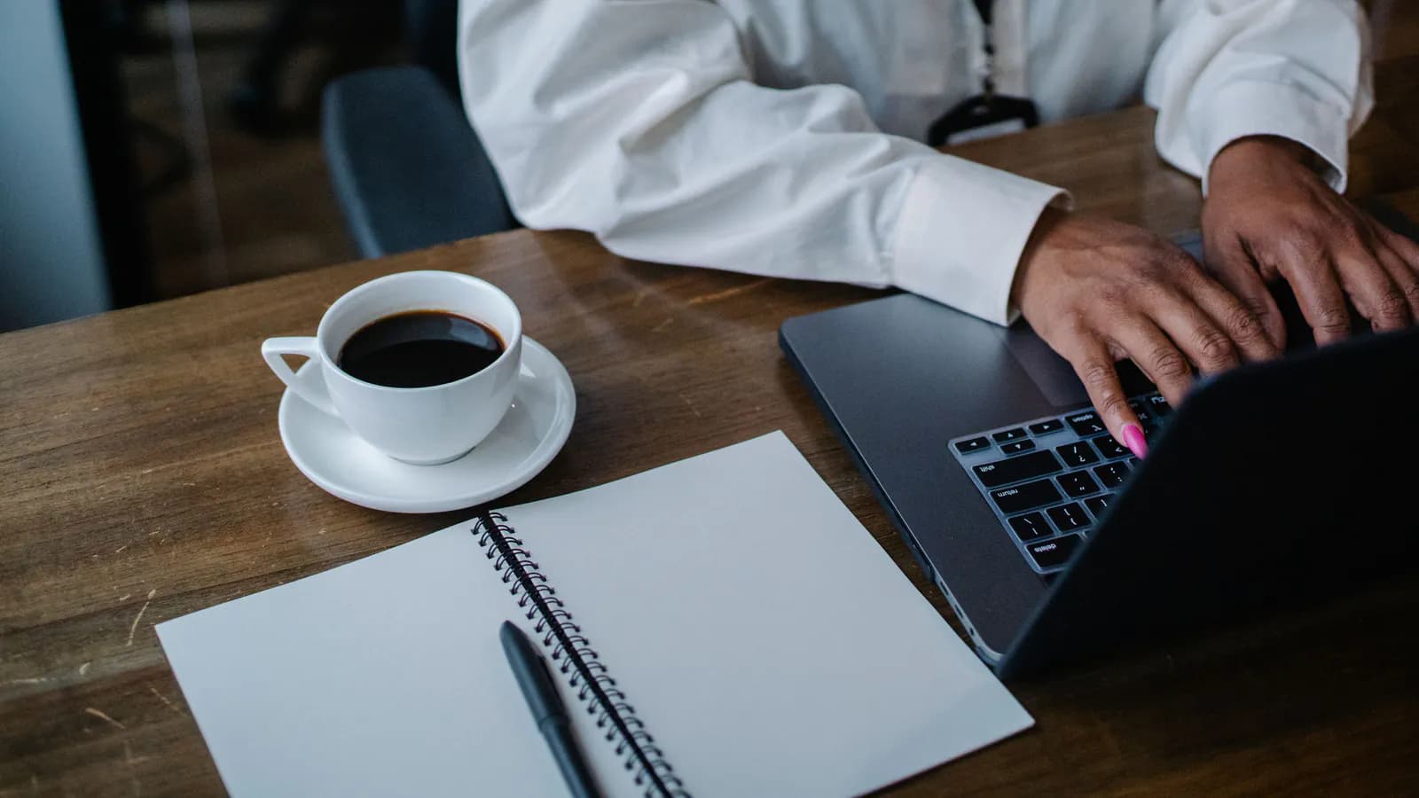 Hands typing on a laptop keyboard on a wooden desk, next to an open notebook and a cup of coffee.