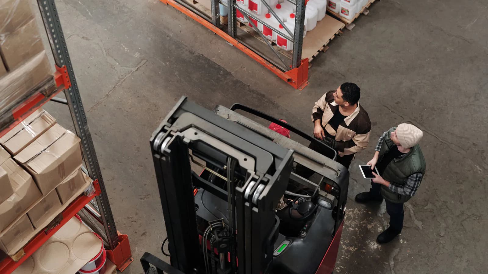 A warehouse worker stands between rows of shelving and scans a barcode on a box with a handheld scanner, stacked inventory boxes visible behind them.