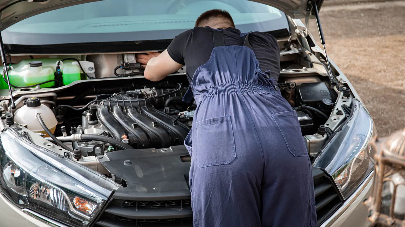 Inside an auto repair garage, a mechanic leans over an open car bonnet checking the engine, with tools hanging on the wall behind him.