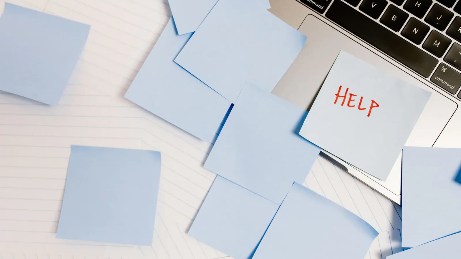 A cluttered desk covered in dozens of blue sticky notes, one reading "HELP" in red marker propped against a laptop keyboard — a mess visibly asking for help.