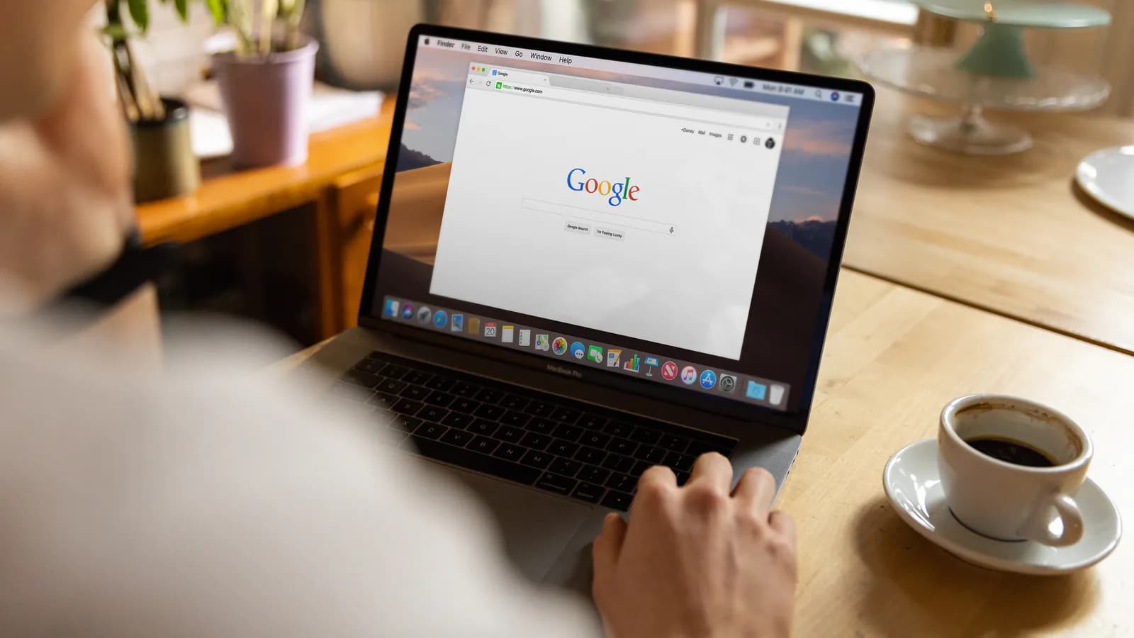 Over-the-shoulder view of someone at a wooden desk, a laptop screen showing a browser open on the Google search homepage, a cup of coffee resting beside it.