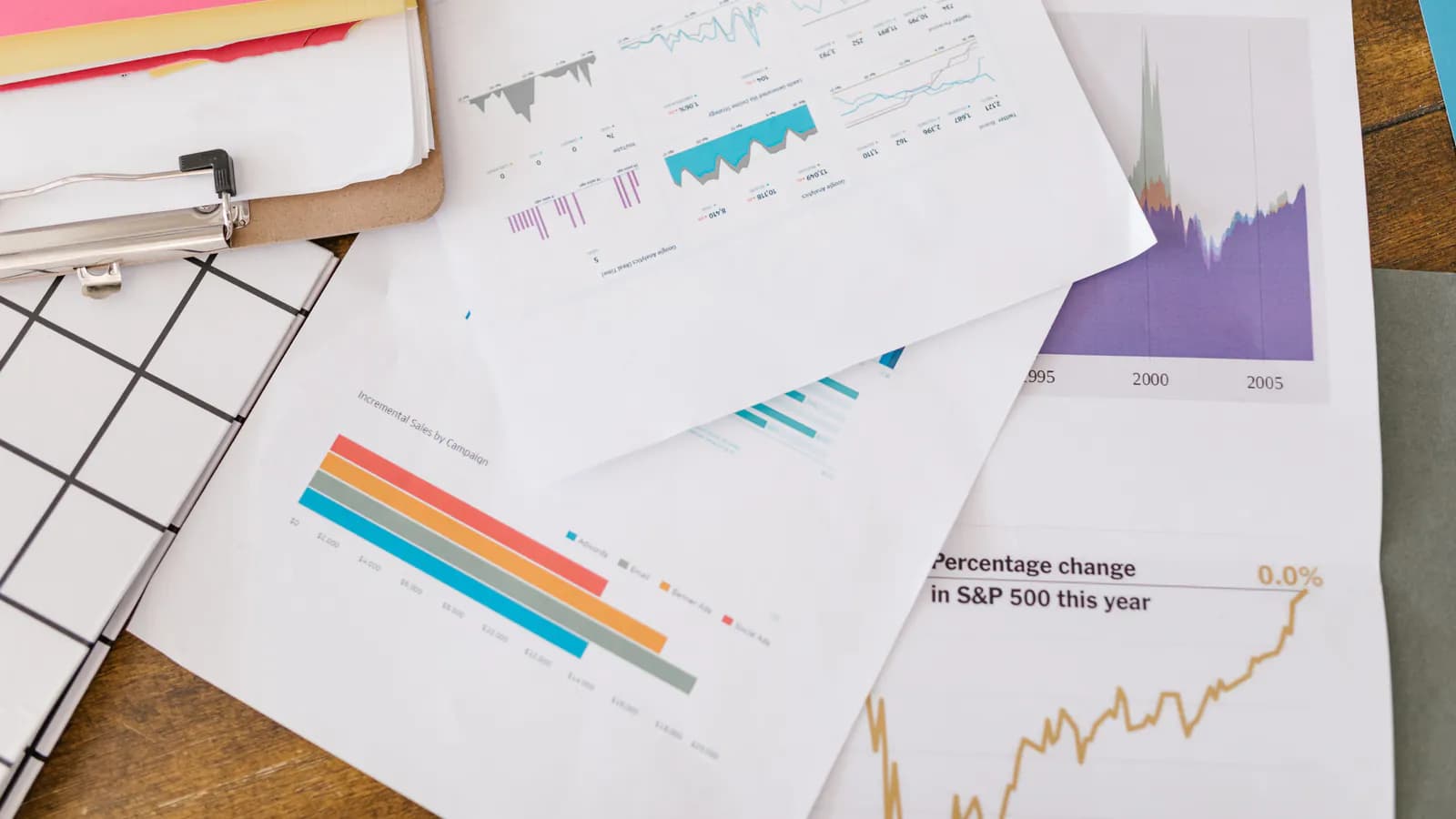 Printed report pages overlapping on a wooden desk — a traffic trend chart, a bar chart of sales by campaign and a percentage-change curve, with a notepad beside them.