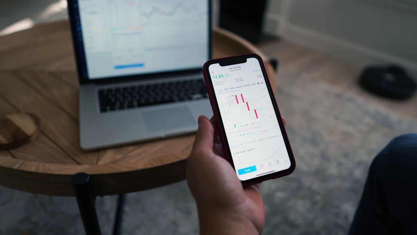 A hand holds a phone with a chart open on screen; behind it on the table sits a laptop showing the same thing.