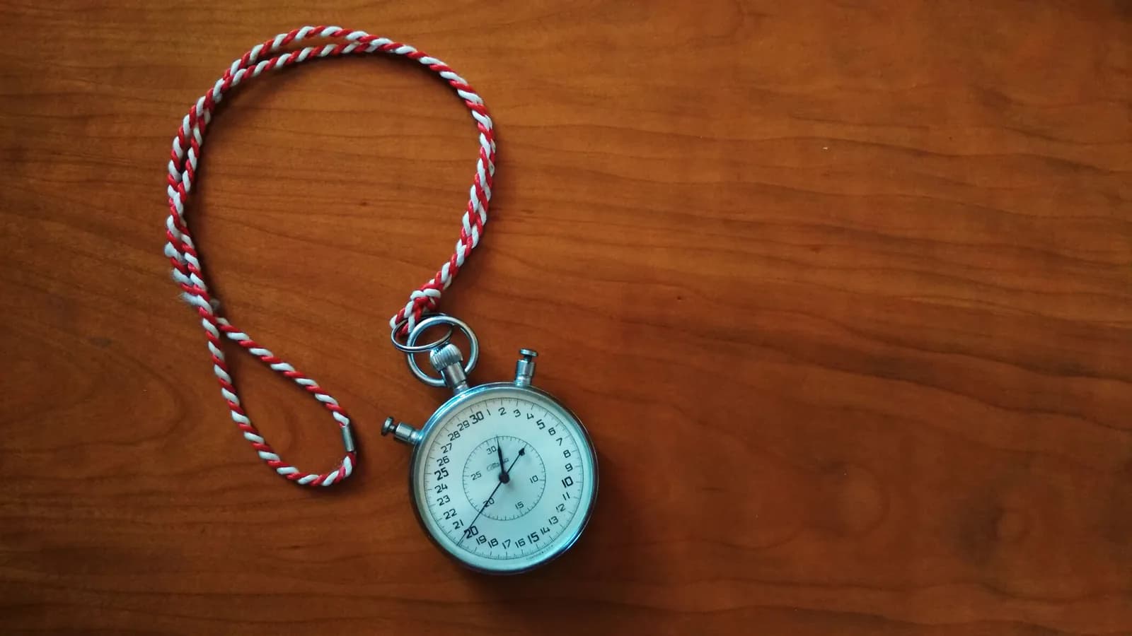 A mechanical stopwatch on a wooden table, its red-and-white cord coiled beside it, the seconds marked from 0 to 30 across the white dial.