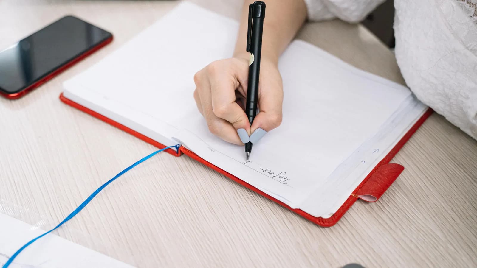 A hand writing with a pen in an open notebook on a white desk, a phone lying beside it.