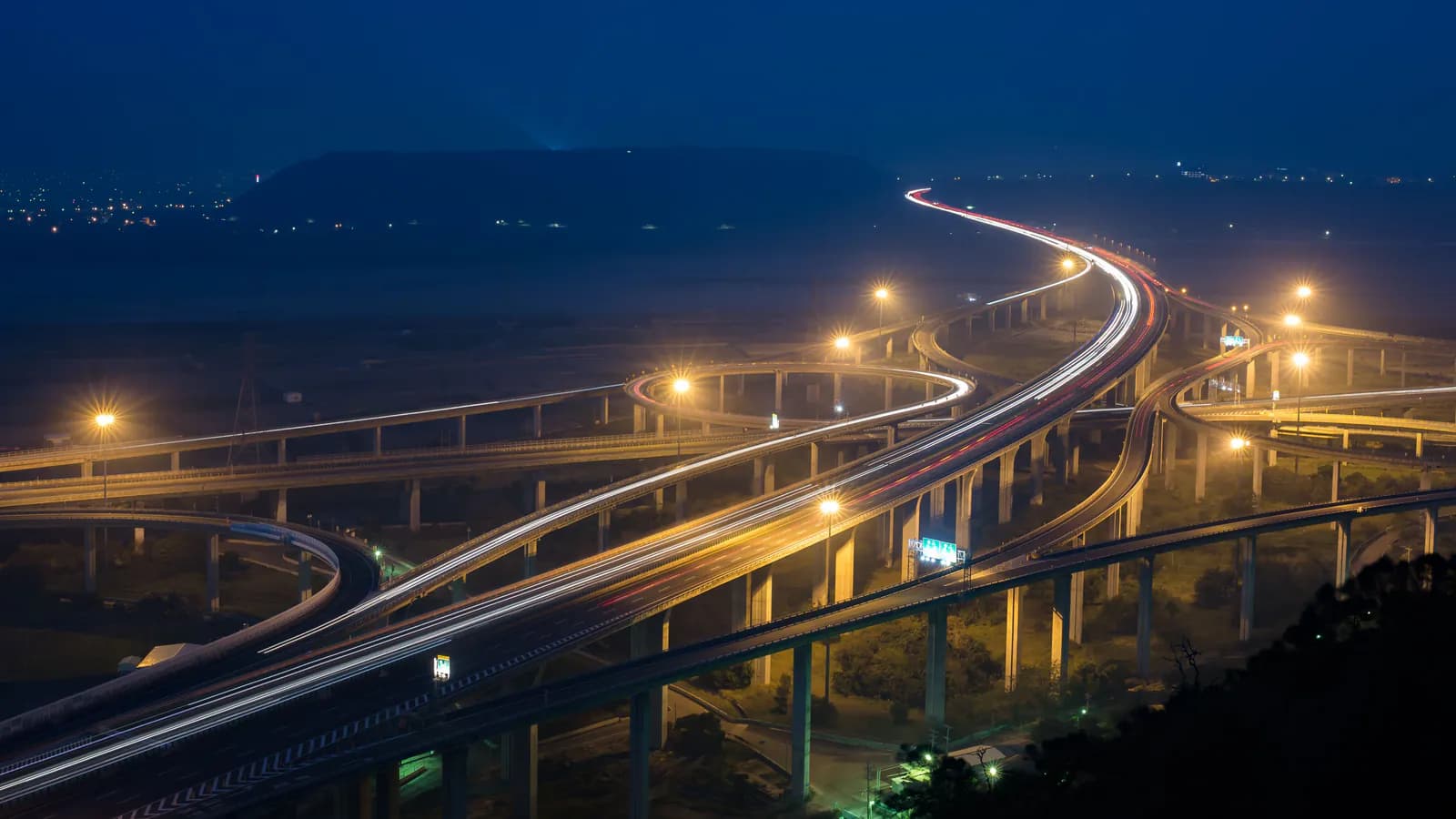 A road interchange at night from above: several flyovers cross and merge into one highway, the car lights drawn out into long white and red streaks.