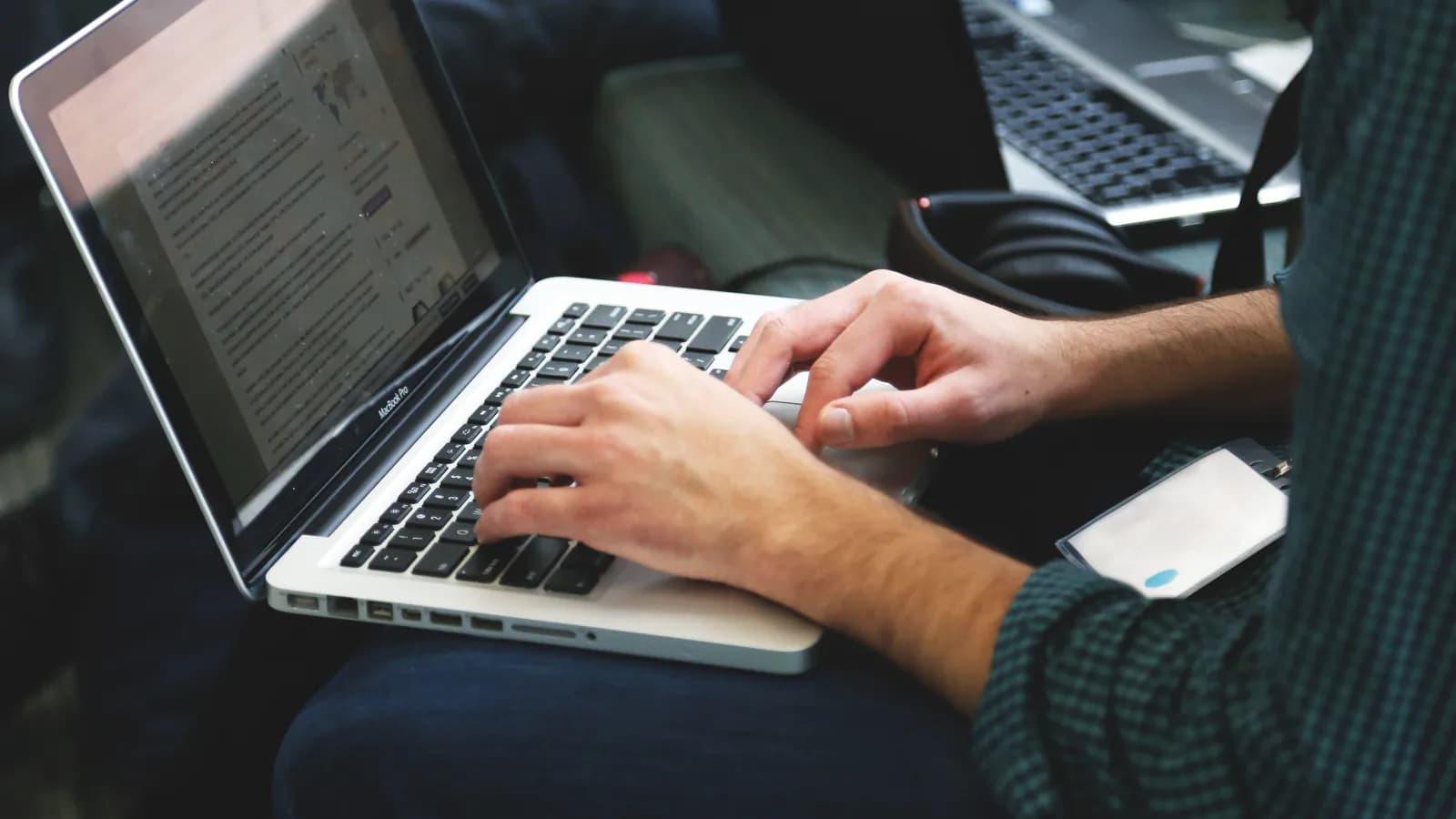 A person types with both hands on a laptop balanced on their knee, a text-filled browser page open on the screen, with a second laptop visible in the background.